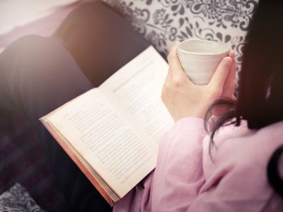 Midsection Of Woman Holding Coffee Cup And Book While Sitting On Sofa At Home