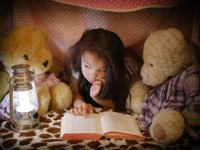 Little girl reading book to teddy bears in fort