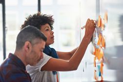 A project manager and team member working at a whiteboard with sticky notes one it