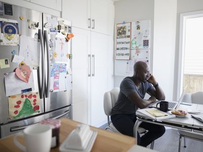 Mature African American man talking on cell phone and working at laptop at kitchen table