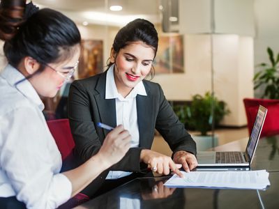A woman in business attire is showing another woman where to sign a document.