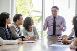 Man standing at conference table gives presentation to colleagues“width=