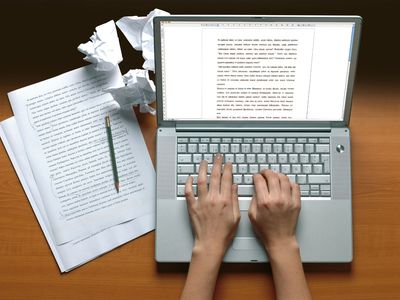 hands typing on a laptop next to printouts and crumbled pieces of paper