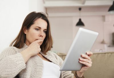 Portrait of middle-aged woman sitting on sofa and working on her tablet computer