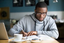 Bespectacled student writing notes before a coffee and laptop